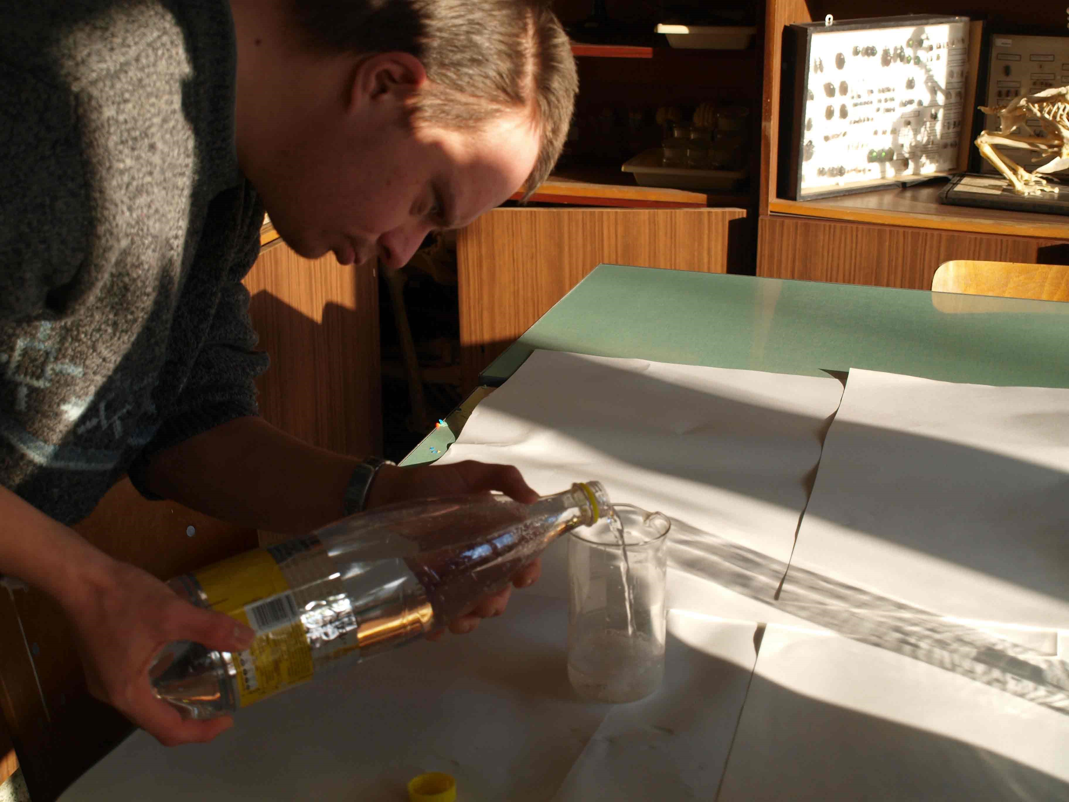 A student pouring a bottle of a soft drink into a small glass.