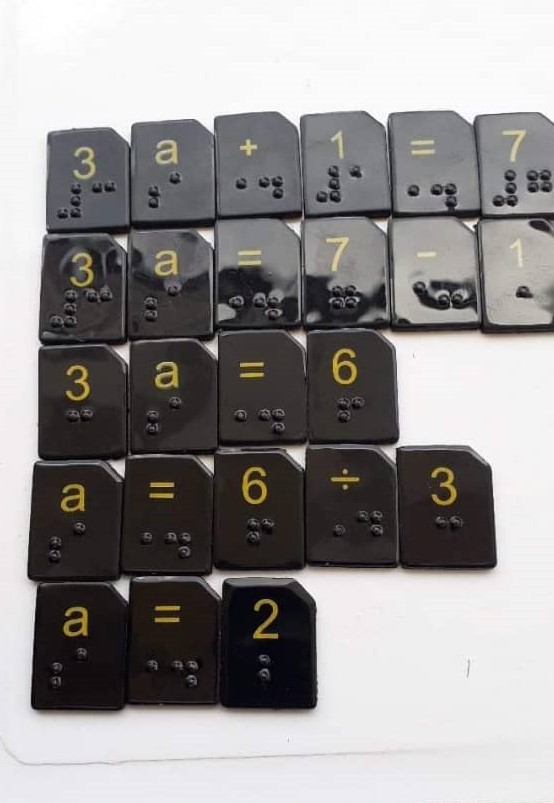 A collection of black tiles with various numbers, letters and symbols printed in yellow with braille cells below.