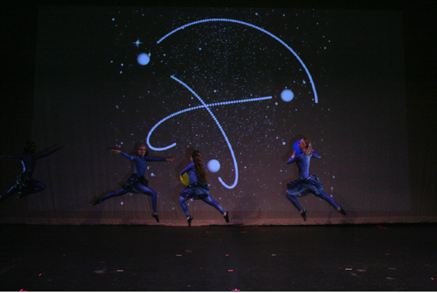 3 people on a stage in dark blue costumes dancing in front of a screen with stars