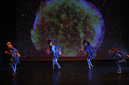 4 people on a stage in dark blue costumes dancing in front of a screen with a star collapsing and exploding as a supernova