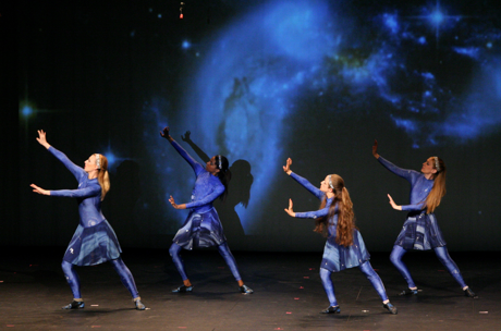 4 people on a stage in dark blue costumes dancing in front of a screen displaying a representation of the collision of galaxy