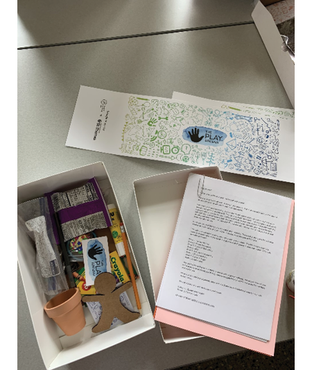 A box containing a set of papers, a clay pot, a cardboard figure, crayola crayons, a glue stick, a pencil, a zip lock bag, a snack, and more items below.